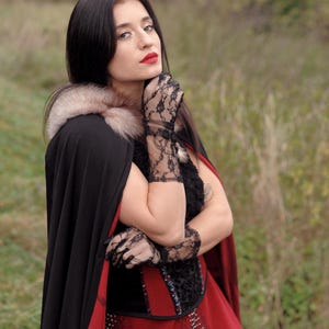 May include: A woman wearing a black cape with a fur collar and a red dress. She is also wearing black lace gloves. The cape has a red lining. The woman has dark hair and red lipstick.