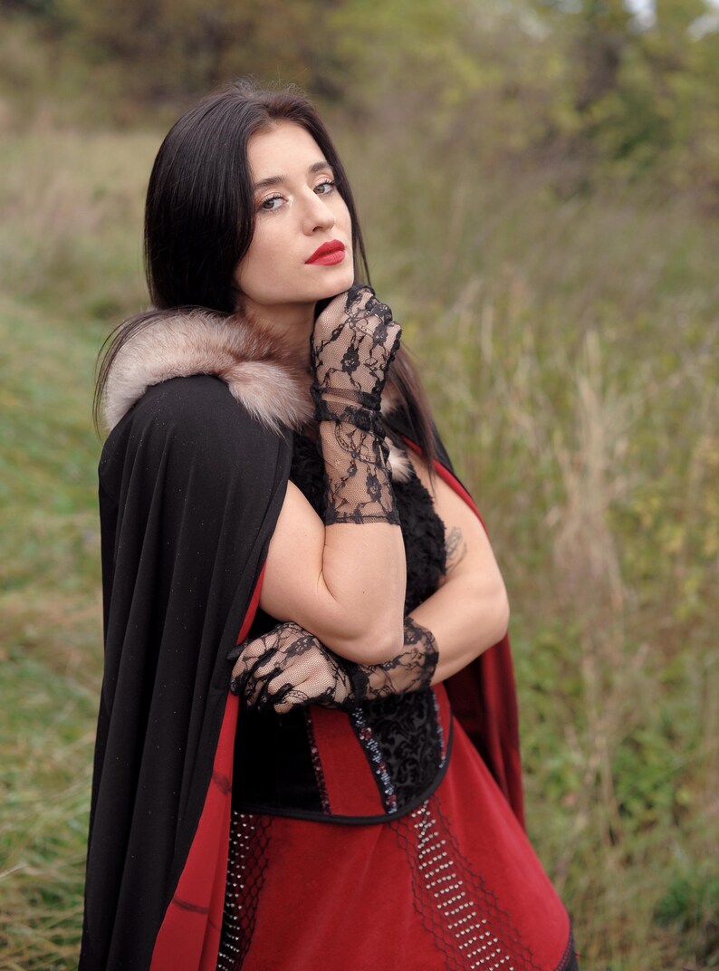 May include: A woman wearing a black cape with a fur collar and a red dress. She is also wearing black lace gloves. The cape has a red lining. The woman has dark hair and red lipstick.