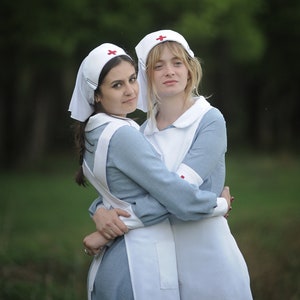 Puede incluir: Dos mujeres con uniformes de enfermera blancos con cruces rojas en sus sombreros. Est&aacute;n de pie juntas y sonriendo. La mujer de la izquierda lleva un vestido azul y la mujer de la derecha lleva un vestido blanco.