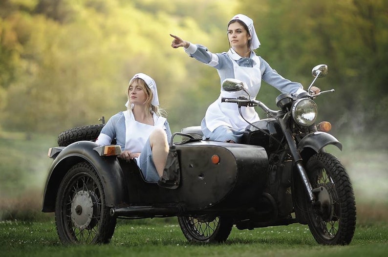 Puede incluir: Dos mujeres vestidas de enfermeras van en una motocicleta negra con sidecar. La mujer en el sidecar mira hacia un lado, mientras que la mujer que conduce la motocicleta apunta hacia la derecha. La motocicleta est&aacute; en un campo de hierba con un fondo borroso de &aacute;rboles.