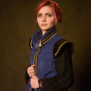 May include: A blue vest with gold trim and black sleeves. The vest has a high collar and a zipper down the front. The sleeves are long and black with lace cuffs. The person in the image has red hair.
