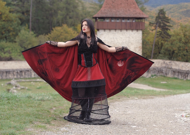 May include: A gothic-style ensemble featuring a red and black cape with a moon design, a black corset, and a red skirt. The outfit includes a black cage skirt and lace gloves, creating a dramatic and theatrical look.