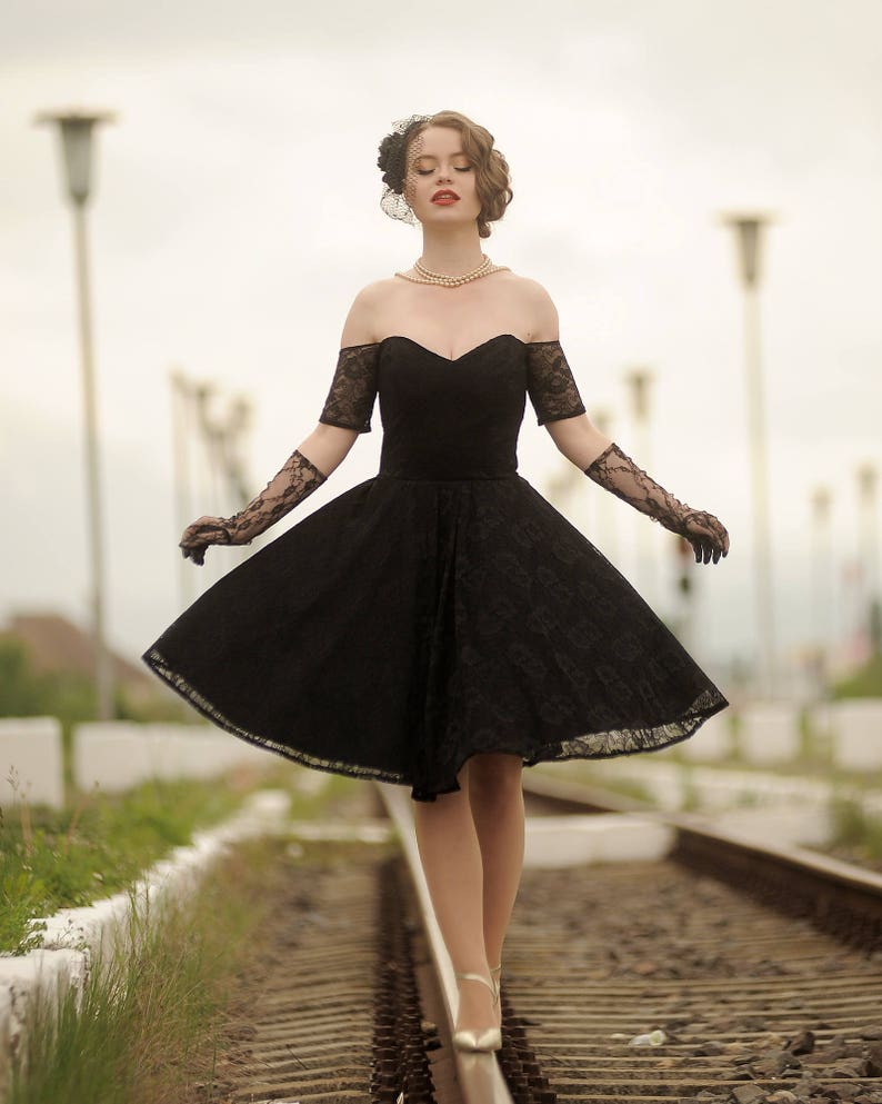 May include: A woman in a black lace dress and gloves walks on a railroad track. She is wearing a pearl necklace and a black veil.
