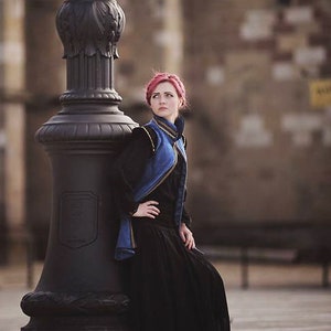 May include: A woman in a black dress and a blue and black jacket leans against a black ornate lamppost. The woman has red hair and is looking off to the side. The background is a blurred view of a building.