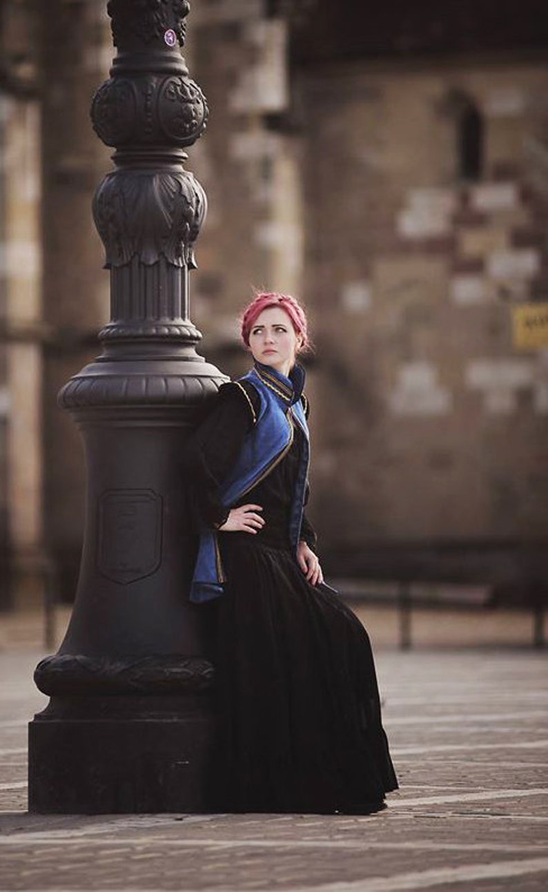 May include: A woman in a black dress and a blue and black jacket leans against a black ornate lamppost. The woman has red hair and is looking off to the side. The background is a blurred view of a building.