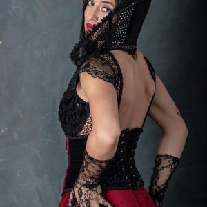 May include: A black corset with lace sleeves and a red skirt. The corset has a detailed floral design and is laced up the back. The outfit includes black lace gloves and a matching hood with a beaded trim. The model is wearing red lipstick.