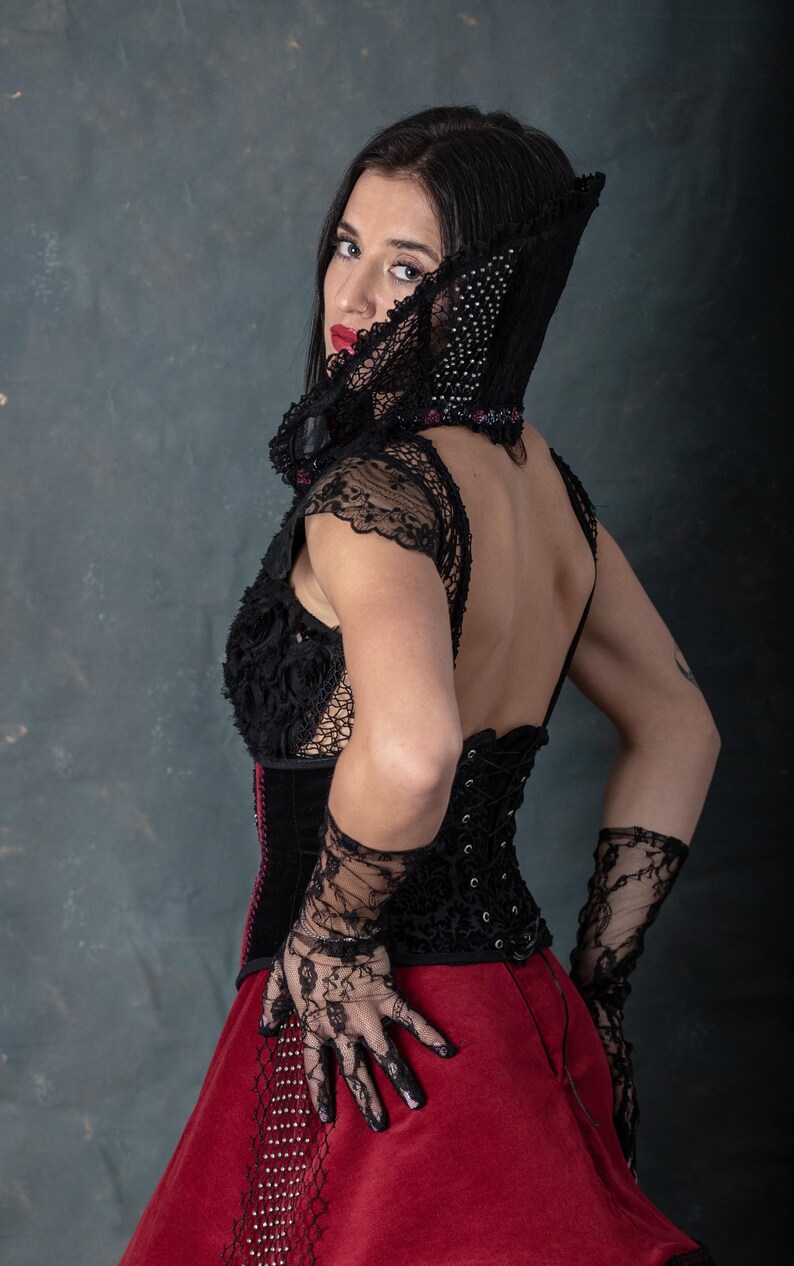 May include: A black corset with lace sleeves and a red skirt. The corset has a detailed floral design and is laced up the back. The outfit includes black lace gloves and a matching hood with a beaded trim. The model is wearing red lipstick.