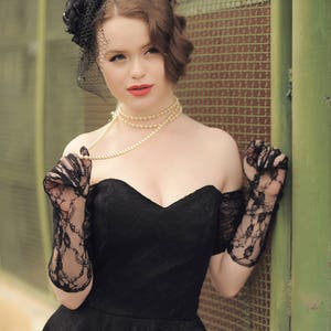 May include: A woman wearing a black off-the-shoulder dress with a lace overlay, black lace gloves, and a pearl necklace. She is wearing a black veil with a flower on top.