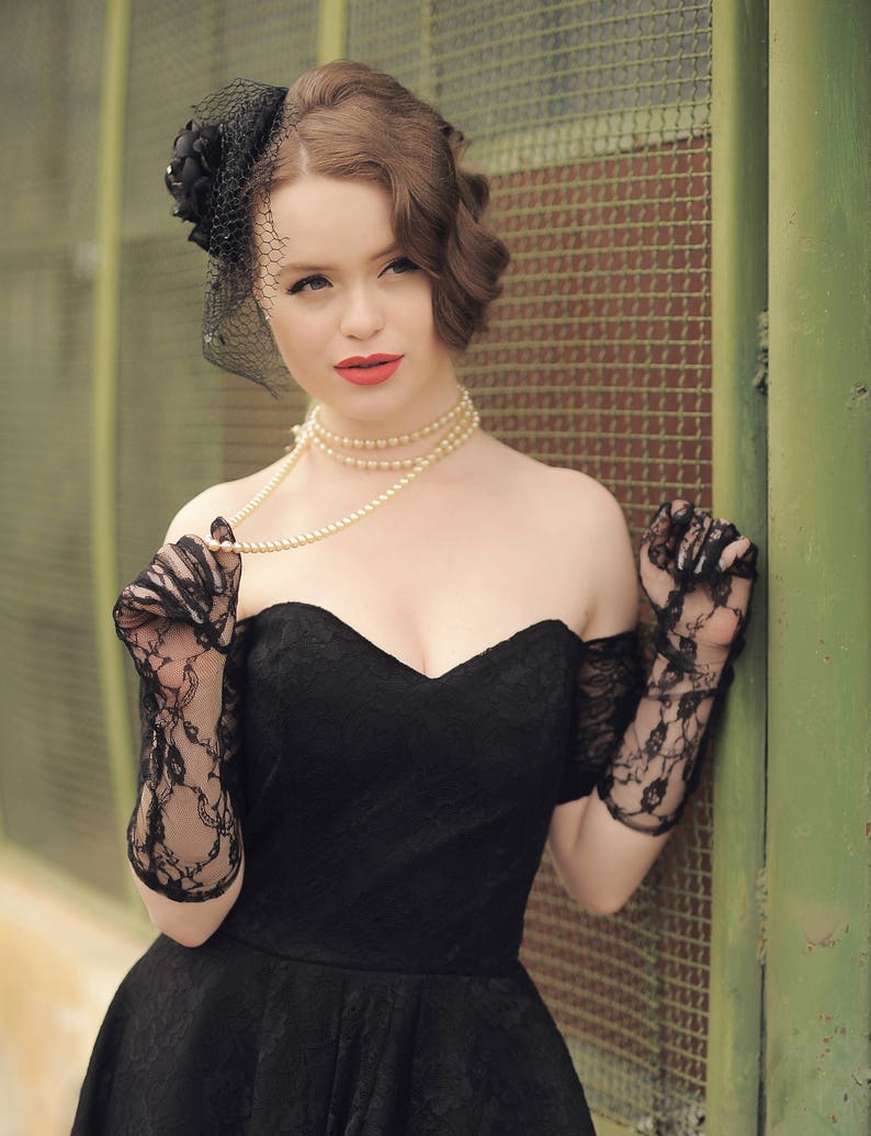 May include: A woman wearing a black off-the-shoulder dress with a lace overlay, black lace gloves, and a pearl necklace. She is wearing a black veil with a flower on top.
