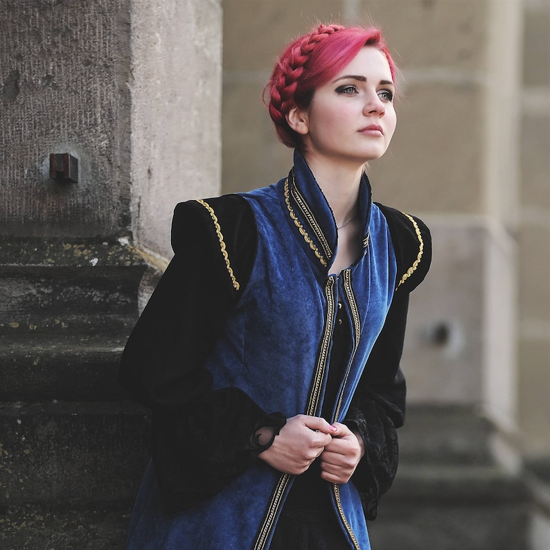 May include: A woman with pink braided hair wearing a blue and black medieval-style vest with gold trim. The vest has a high collar and is buttoned up the front. The sleeves are black and the background is a stone wall.
