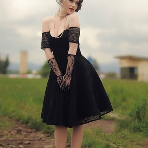 May include: A woman in a black lace dress with a black hat and veil. She is wearing pearl jewelry and black lace gloves. She is standing on a path with green grass and rocks.