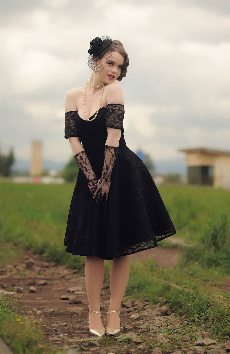 May include: A woman in a black lace dress with a black hat and veil. She is wearing pearl jewelry and black lace gloves. She is standing on a path with green grass and rocks.