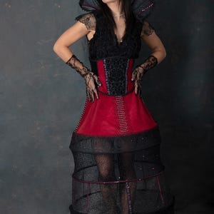 May include: A dramatic black and red costume with a high collar and a structured skirt. The bodice is black with lace detailing and a red panel. The skirt has a cage-like structure, and the overall design is theatrical.