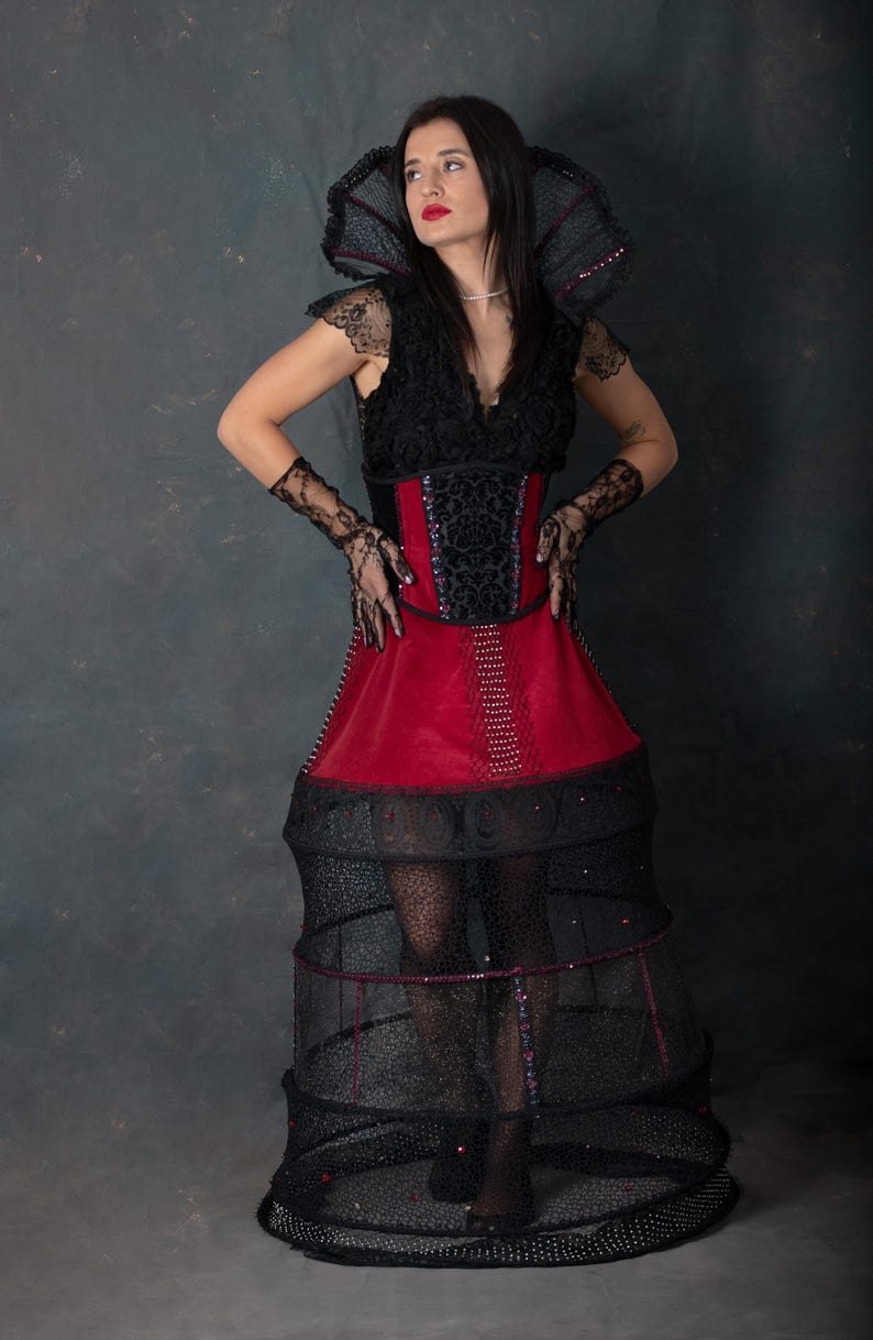 May include: A dramatic black and red costume with a high collar and a structured skirt. The bodice is black with lace detailing and a red panel. The skirt has a cage-like structure, and the overall design is theatrical.