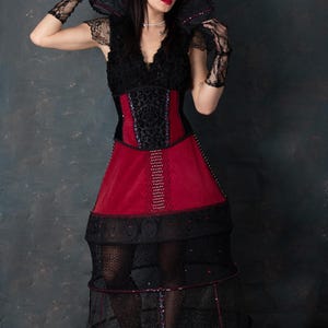 May include: A gothic-style dress with a black lace bodice and a red velvet skirt. The dress features a structured, cage-like skirt and matching lace gloves. The model is wearing a pearl necklace and has dark hair and red lipstick.