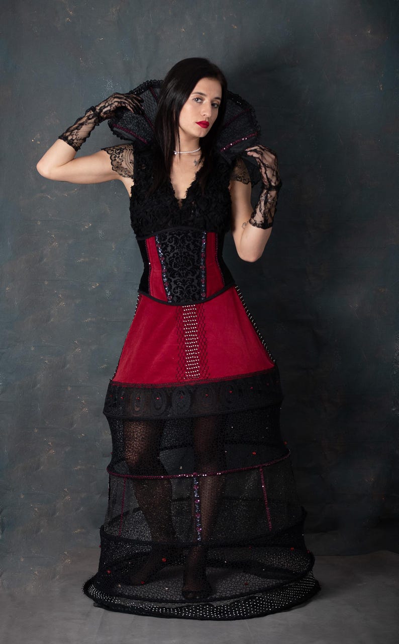 May include: A gothic-style dress with a black lace bodice and a red velvet skirt. The dress features a structured, cage-like skirt and matching lace gloves. The model is wearing a pearl necklace and has dark hair and red lipstick.