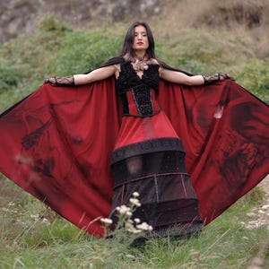 May include: A gothic-style dress with a black bodice and a red skirt. The dress features a black cape with a dark print. The model is standing in a grassy area with arms outstretched, showcasing the dress's design.