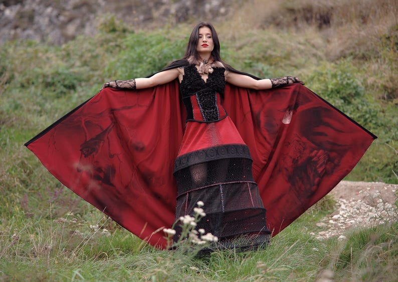 May include: A gothic-style dress with a black bodice and a red skirt. The dress features a black cape with a dark print. The model is standing in a grassy area with arms outstretched, showcasing the dress's design.