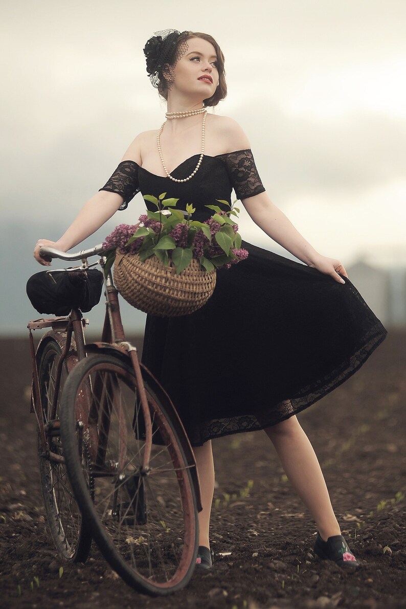 May include: A woman in a black lace dress and a pearl necklace rides a vintage bicycle with a wicker basket filled with purple flowers.