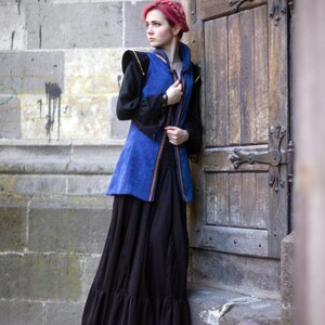 May include: A woman wearing a medieval-style outfit stands by a wooden door. The outfit includes a blue vest with gold trim, a black long-sleeved shirt, and a long black skirt. The woman has red hair.