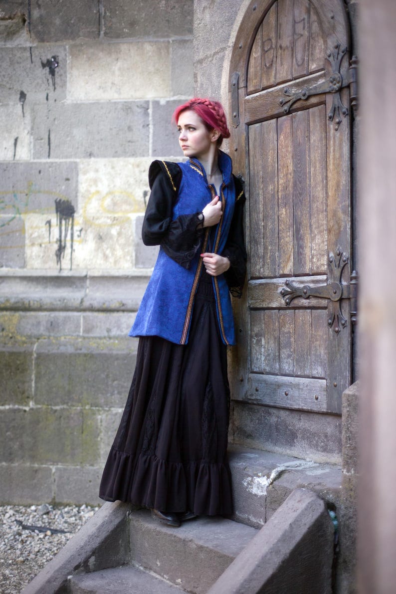 May include: A woman wearing a medieval-style outfit stands by a wooden door. The outfit includes a blue vest with gold trim, a black long-sleeved shirt, and a long black skirt. The woman has red hair.