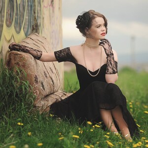 May include: A woman in a black lace dress with a pearl necklace and a black hat sits on a couch in front of a graffiti wall.