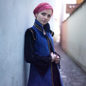 May include: A woman with pink hair wearing a blue and black vest with gold trim, over a black long-sleeved top. The vest has a high collar and is buttoned down the front. The woman is standing in a narrow alleyway, leaning against a light blue wall.