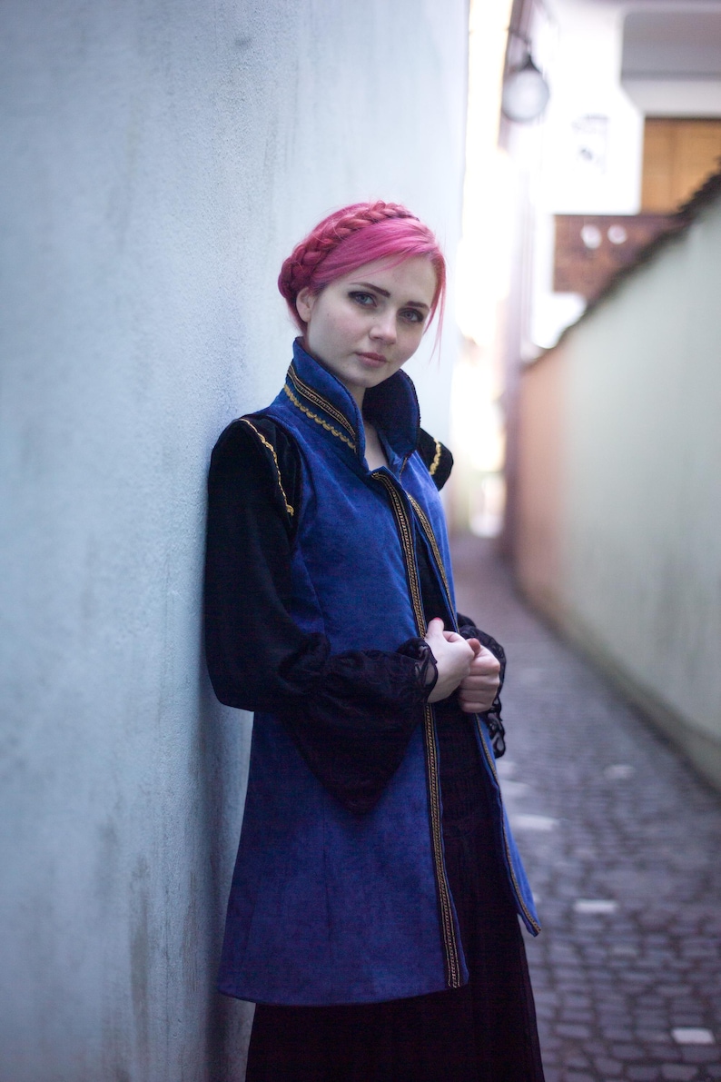 May include: A woman with pink hair wearing a blue and black vest with gold trim, over a black long-sleeved top. The vest has a high collar and is buttoned down the front. The woman is standing in a narrow alleyway, leaning against a light blue wall.