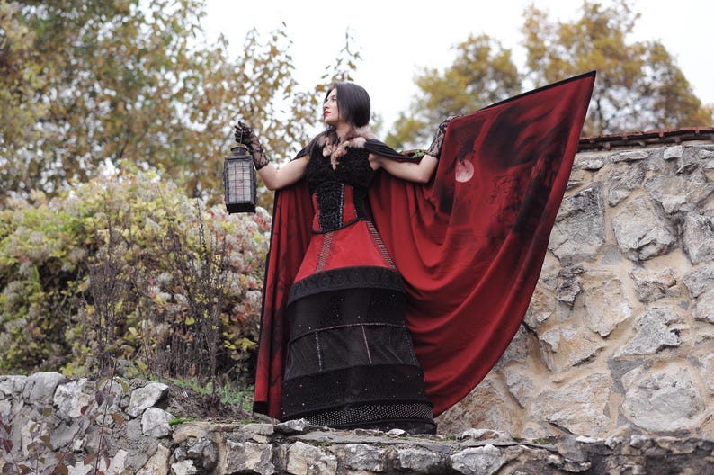 May include: A woman in a gothic-style costume stands on a stone wall, holding a lantern. The outfit features a red and black dress with a matching cape. The cape has a dark design and is spread out, creating a dramatic effect.