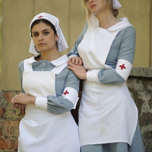 Puede incluir: Dos mujeres con uniformes de enfermera azules y blancos con cruces rojas en las mangas. Est&aacute;n de pie junto a una pared de ladrillo.