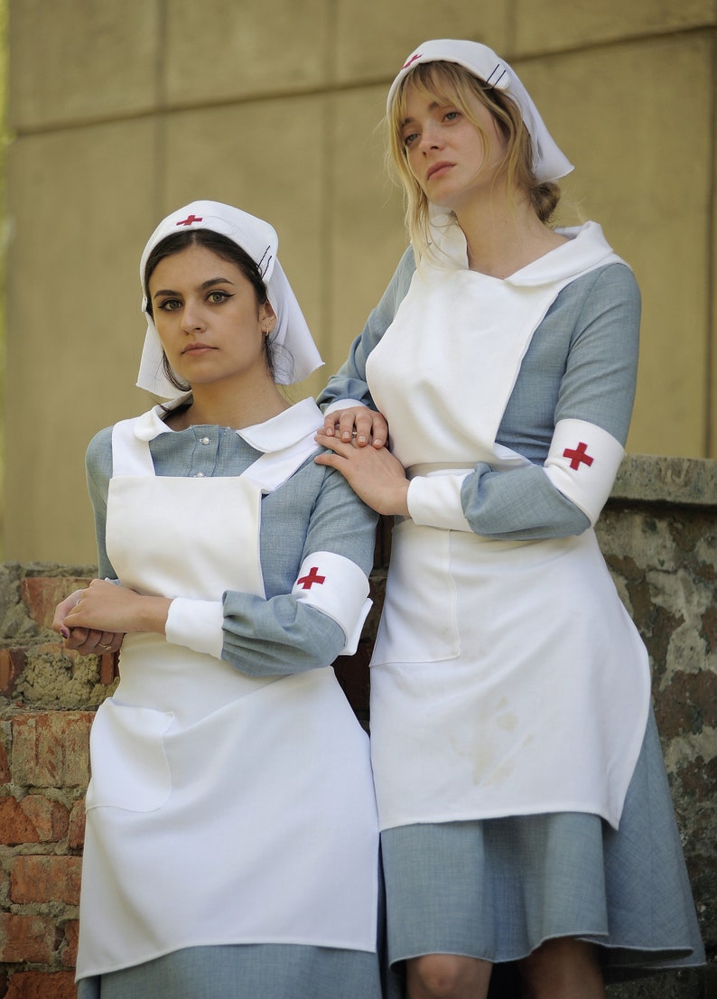 Puede incluir: Dos mujeres con uniformes de enfermera azules y blancos con cruces rojas en las mangas. Est&aacute;n de pie junto a una pared de ladrillo.