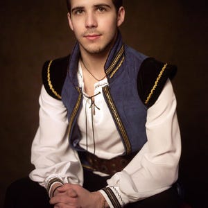 May include: A person wearing a white shirt with a lace-up neckline, a blue vest with gold trim, and black accents. The vest has black shoulder pads with gold trim. The person is wearing a brown belt and black pants.
