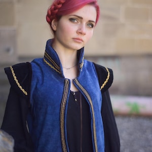 May include: A blue corduroy vest with gold trim and black accents. The vest has a high collar and is worn over a black long-sleeved top. The outfit has a medieval or renaissance theme.