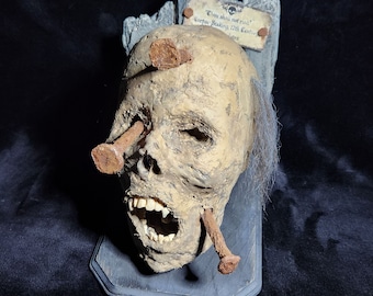 Può includere: Una scultura di teschio umano invecchiato con chiodi arrugginiti conficcati negli occhi e nella testa, montata su una placca di legno grigio. Un piccolo cartello sopra recita "Thou shall not rise!" e "Corpse Staking, 17th Century". Il teschio ha denti scoperti e capelli grigi.