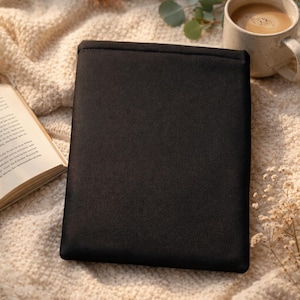 May include: A black book cover made of a smooth, matte fabric. The cover is rectangular and appears to be empty. The cover is resting on a cream-colored, textured surface, with a cup of coffee and an open book in the background.