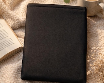 May include: A black book cover made of a smooth, matte fabric. The cover is rectangular and appears to be empty. The cover is resting on a cream-colored, textured surface, with a cup of coffee and an open book in the background.