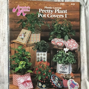 May include: A magazine cover by Annie's Attic, titled "Pretty Plant Pot Covers I", showcasing plastic canvas pot covers with plants. Designs include a house, a heart, and geometric patterns. The background is a wooden surface.