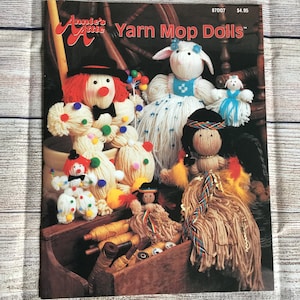 May include: A vintage book cover titled "Annie's Attic Yarn Mop Dolls" featuring various yarn dolls. The dolls include a clown, a sheep, and a Native American doll. The cover is set against a wooden background.