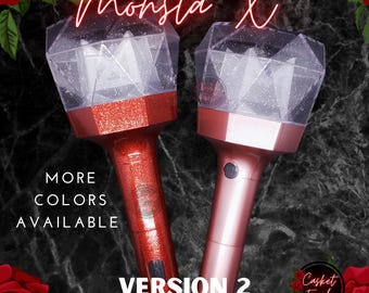 May include: Two Monsta X Version 2 lightsticks with clear, faceted, diamond-shaped tops containing internal structures. One handle is a glittery red, and the other is a smooth, metallic rose gold. Text reads Monsta X, More Colors Available, and Version 2 Lightstick Skins.