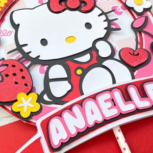 Puede incluir: Un topper de pastel Hello Kitty rosa y rojo con un fondo blanco. El topper presenta un personaje de Hello Kitty con un lazo rojo y un vestido rojo. Las palabras "Happy Birthday" están escritas en letras amarillas alrededor de la parte superior del topper. El nombre "Anaelle" está escrito en letras blancas en una pancarta roja en la parte inferior del topper.