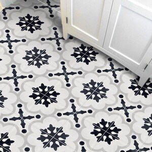 May include: Close-up of patterned floor tiles with a repeating design. The tiles display black snowflake-like shapes and cross patterns on a light grey and white background. A white cabinet is visible in the background.