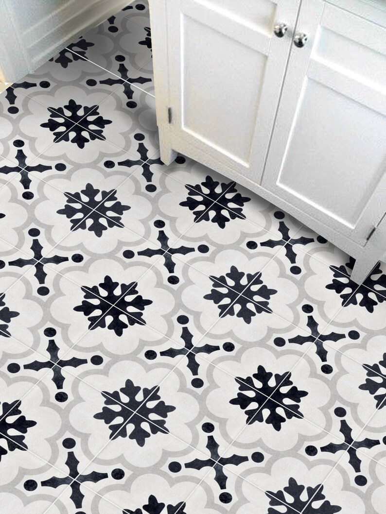May include: Close-up of patterned floor tiles with a repeating design. The tiles display black snowflake-like shapes and cross patterns on a light grey and white background. A white cabinet is visible in the background.