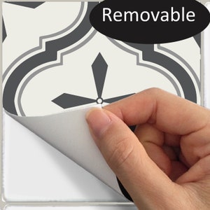 May include: A close-up of a decorative tile with a geometric pattern in shades of gray and cream. The tile is labeled "Removable" in a black oval. A hand is peeling back a corner of the tile, revealing a white adhesive backing.