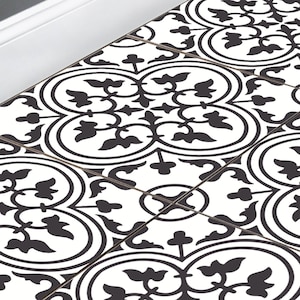 May include: Square tiles with a black and white floral pattern. The design features a repeating motif of stylized flowers and leaves. The tiles are bordered in a dark colour and are arranged in a grid.