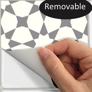 Puede incluir: Una baldosa cuadrada con un patr&oacute;n geom&eacute;trico en gris y blanco roto se est&aacute; despegando, revelando una superficie blanca. La palabra "Removable" est&aacute; escrita en blanco sobre un &oacute;valo negro. Una mano est&aacute; despegando la baldosa.