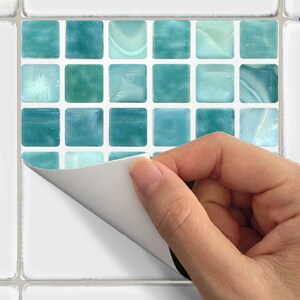 May include: A sheet of peel-and-stick mosaic tiles with a grid of small, square tiles in shades of teal and aqua. A hand is peeling back the backing paper, revealing the adhesive. The tiles are designed for easy application.