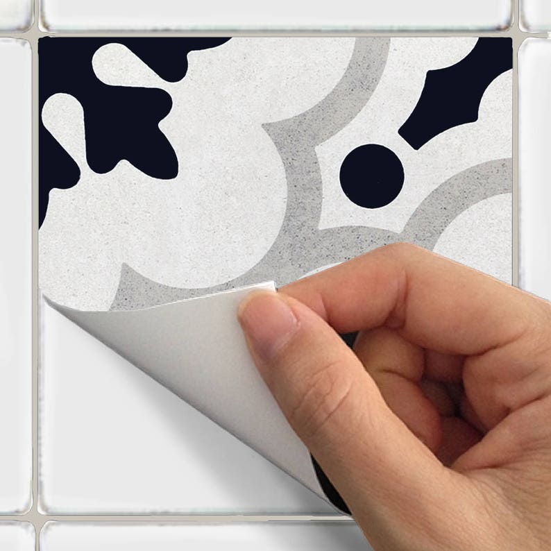 May include: A decorative tile sticker featuring a repeating floral pattern in white, grey, and navy blue. The sticker is being peeled back to reveal the adhesive backing. The design has a textured appearance, suitable for home decor projects.