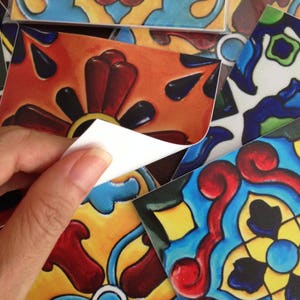 Puede incluir: Una mano quitando una pegatina de una baldosa colorida. La baldosa presenta un dise&ntilde;o floral con detalles rojos, naranjas, amarillos y negros. Se ven otras baldosas con patrones similares en azul, amarillo y rojo.