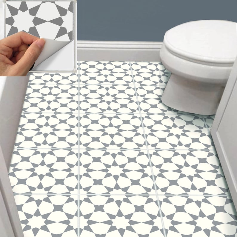 Puede incluir: Azulejos de suelo con dise&ntilde;o geom&eacute;trico en un ba&ntilde;o. Los azulejos presentan un dise&ntilde;o repetitivo en forma de estrella en gris y blanco. Una mano despega una baldosa de una muestra, mostrando el adhesivo. Hay un inodoro blanco.