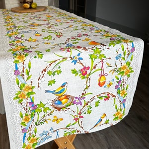 以下が含まれることがあります： 白いテーブルランナーに、花と鳥の模様が施されています。模様には青い鳥、黄色の花、緑の葉が描かれています。ランナーは白いレースの縁取りがされています。